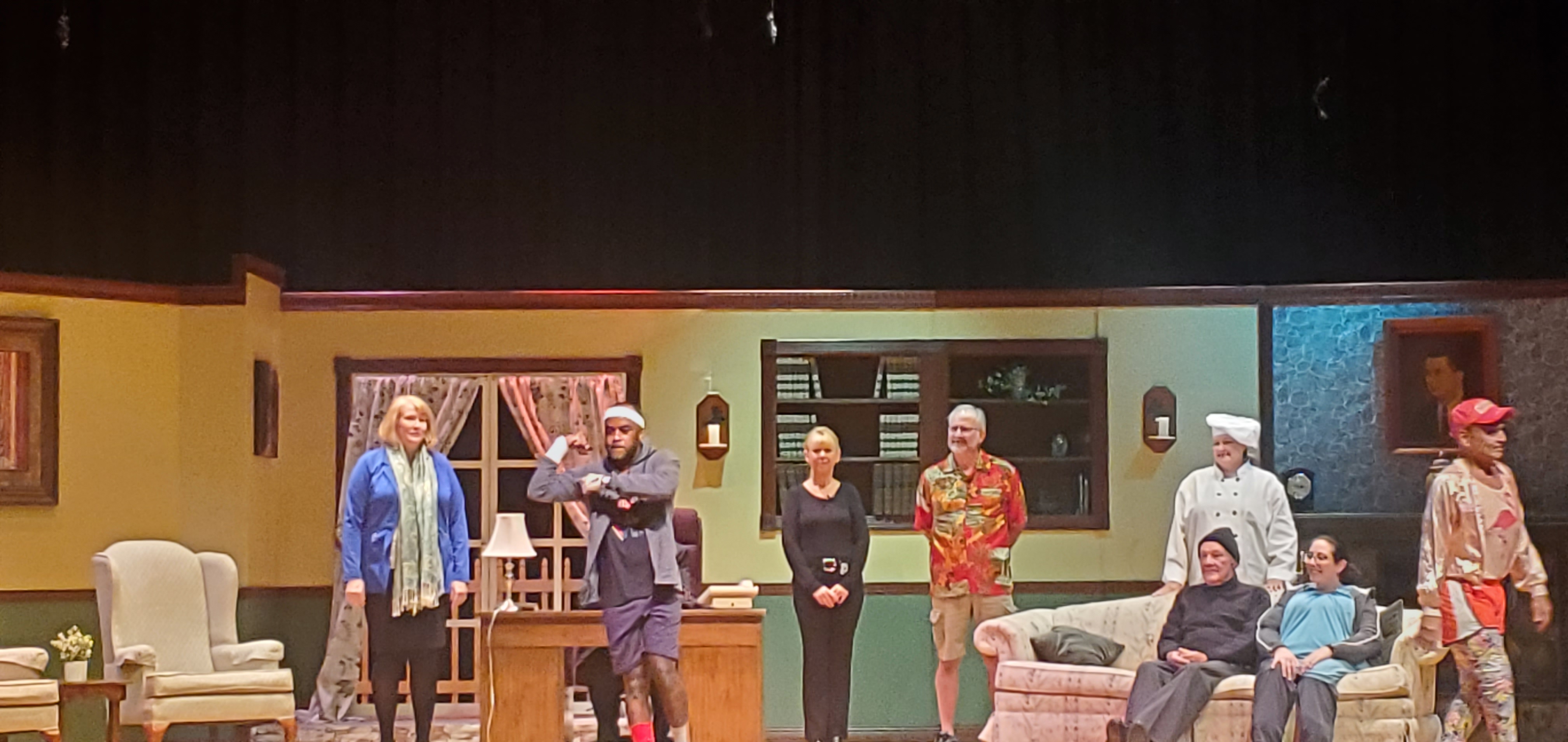 From left to right: a woman in a blue jacket and light green scarf; a man in a black suit sitting behind a desk; a man wearing a white headband, purple shorts and a gray jacket standing in front of the desk; a woman in a black top and pants;
       a man in a red Hawaiian shirt and brown shorts; a man in a black pullover, gray pants and black cap and a woman in a long bluish green top sitting on a couch; a chef standing behind the couch; and a man in an orange running outfit.