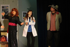 The man in the black dress and red sash, the woman doctor in the labcoat, and the man in the jacket and large hat